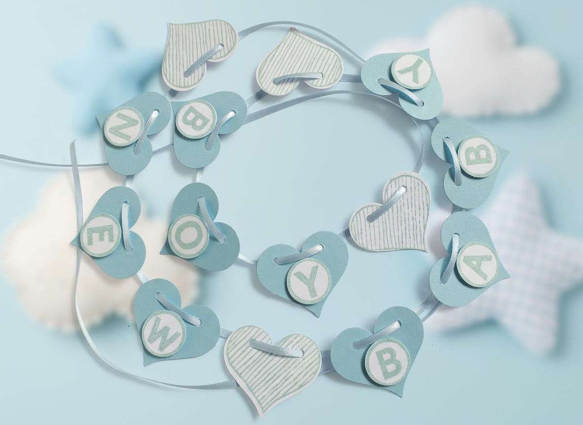 A hand-crafted banner for a baby boy, featuring blue and white hearts and letters spelling 'NEW BABY BOY' on a satin ribbon.