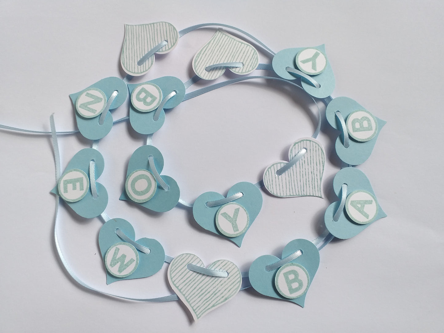 A hand-crafted banner for a baby boy, featuring blue and white hearts and letters spelling 'NEW BABY BOY' on a satin ribbon.