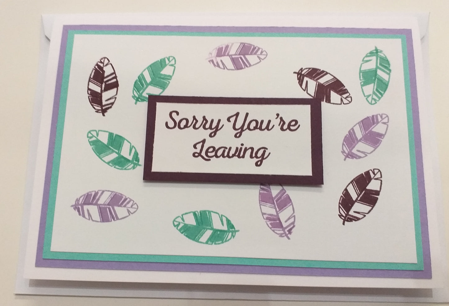 A greeting card with a white background and a message 'Sorry You're Leaving' accompanied by a graphic of feathers in various shades of green and purple.