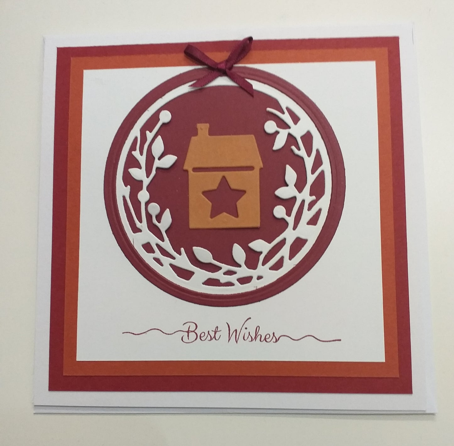 A handmade greeting card with a house and star cut-out design, titled 'Best Wishes' on the front, and a red and orange color scheme. It includes an envelope.