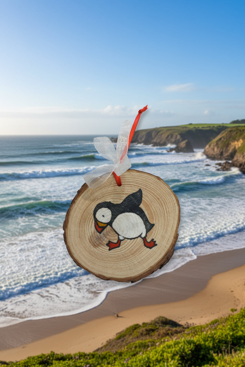 A wood slice decoration featuring a colorful illustration of a puffin, with a red ribbon hanging on the top.