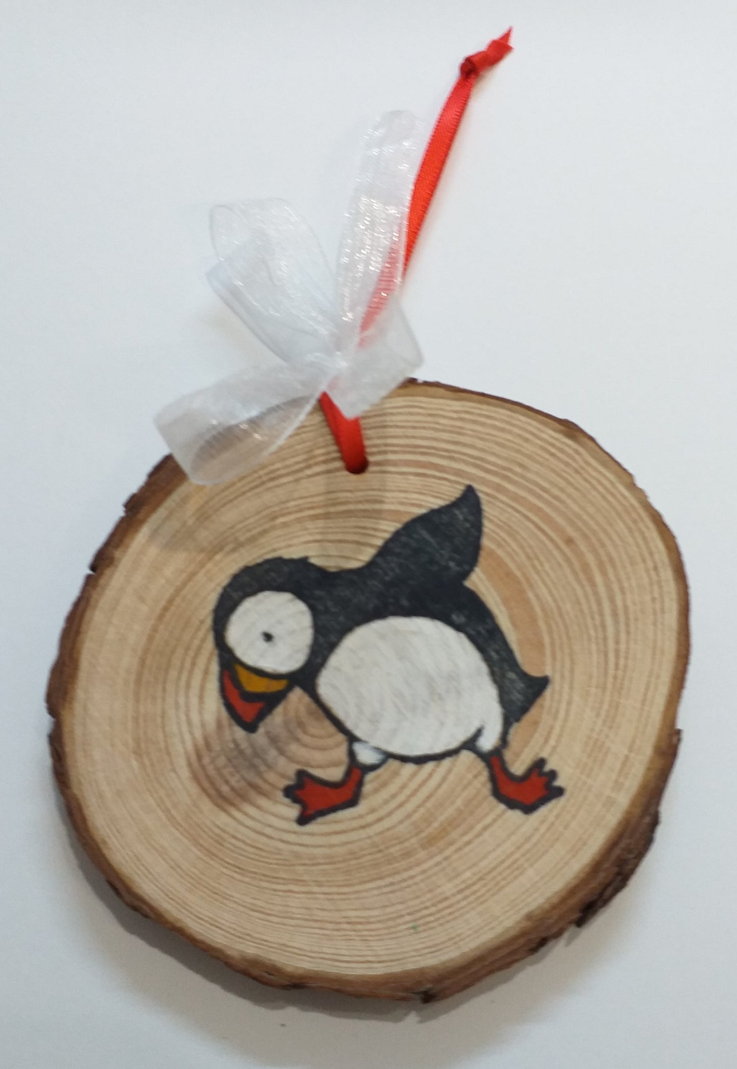 A wood slice decoration featuring a colorful illustration of a puffin, with a red ribbon hanging on the top.