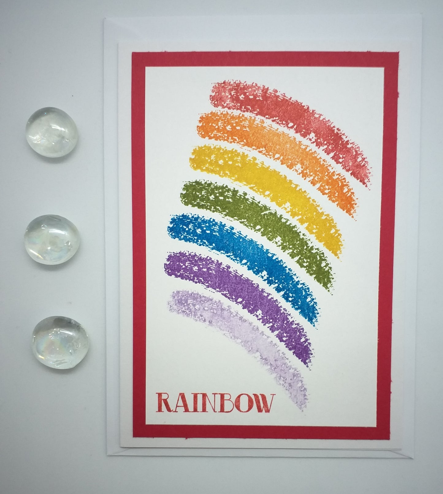 A colorful hand-stamped greeting card with a rainbow design on a red background, with a white border and text label 'RAINBOW'.