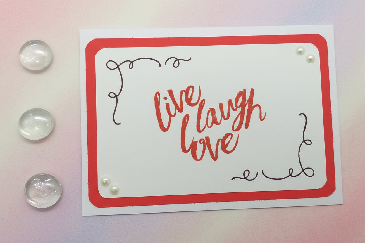 A greeting card with a red and white color scheme, featuring hand-stamped images and pearl embellishments, with the phrase 'Live Laugh Love' written in a cursive script.