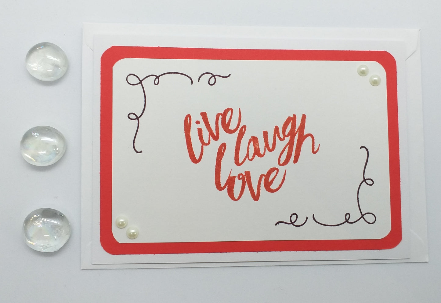 A greeting card with a red and white color scheme, featuring hand-stamped images and pearl embellishments, with the phrase 'Live Laugh Love' written in a cursive script.