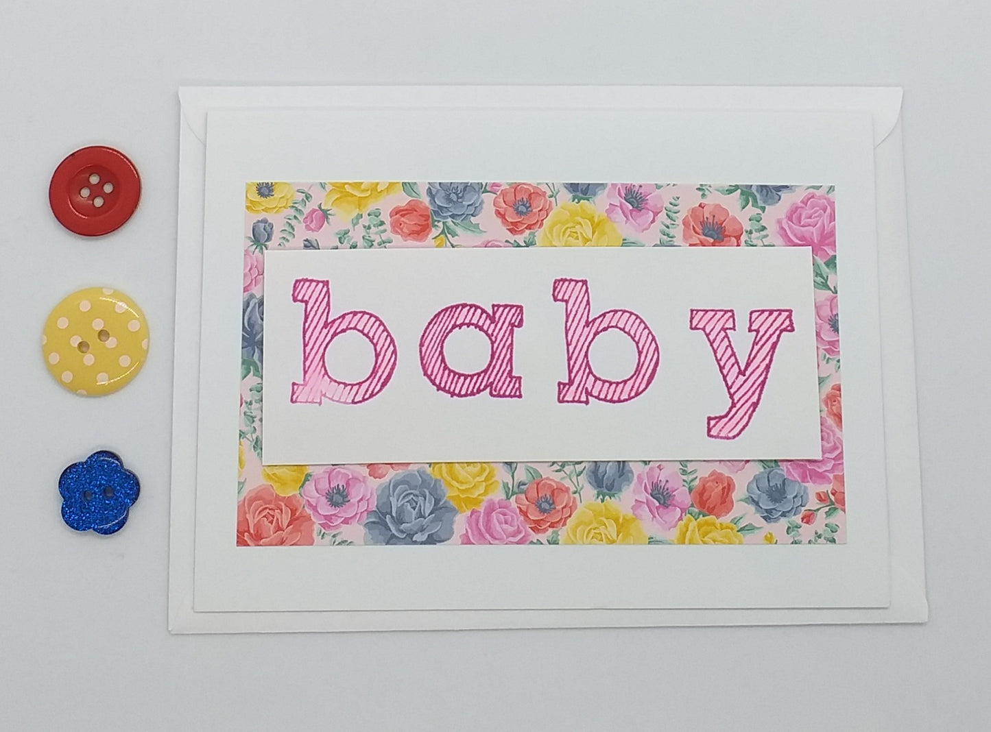 A hand stamped baby card with the word 'baby' in the center, presented on a white background with an envelope and colorful buttons to the side.
