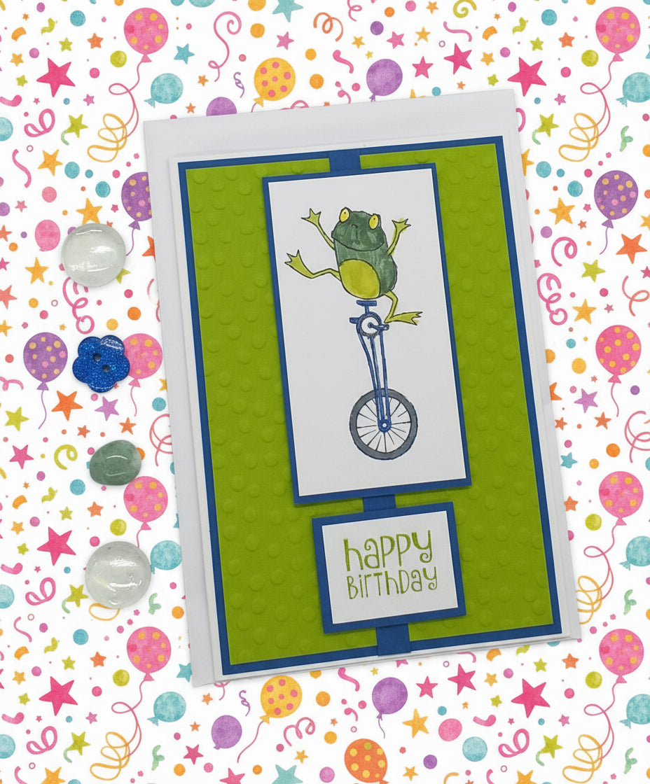 A handmade, multi-layered birthday card featuring a colorful frog riding a bicycle on a green background, accompanied by an envelope and a small glass bead ornament.