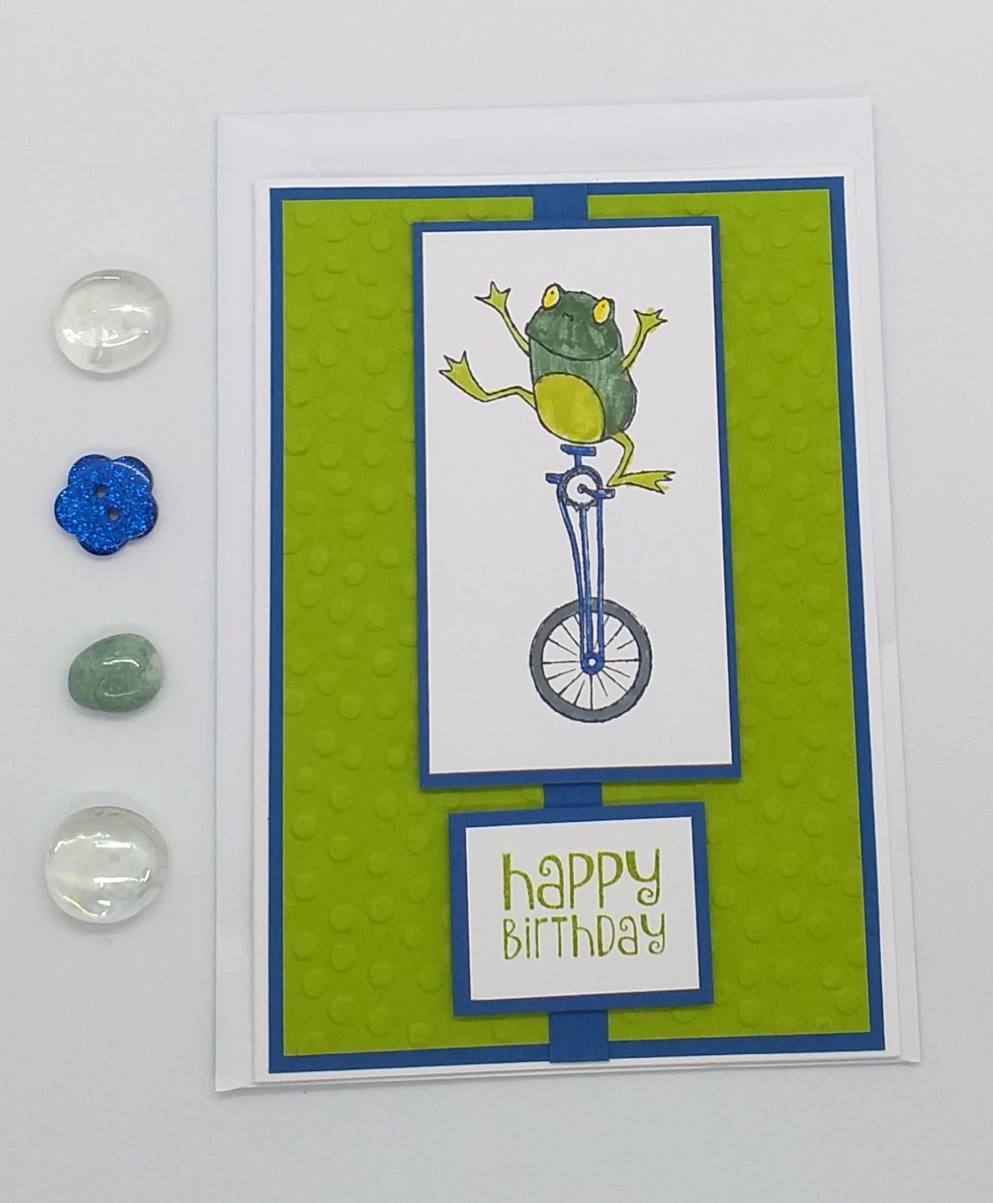 A handmade, multi-layered birthday card featuring a colorful frog riding a bicycle on a green background, accompanied by an envelope and a small glass bead ornament.