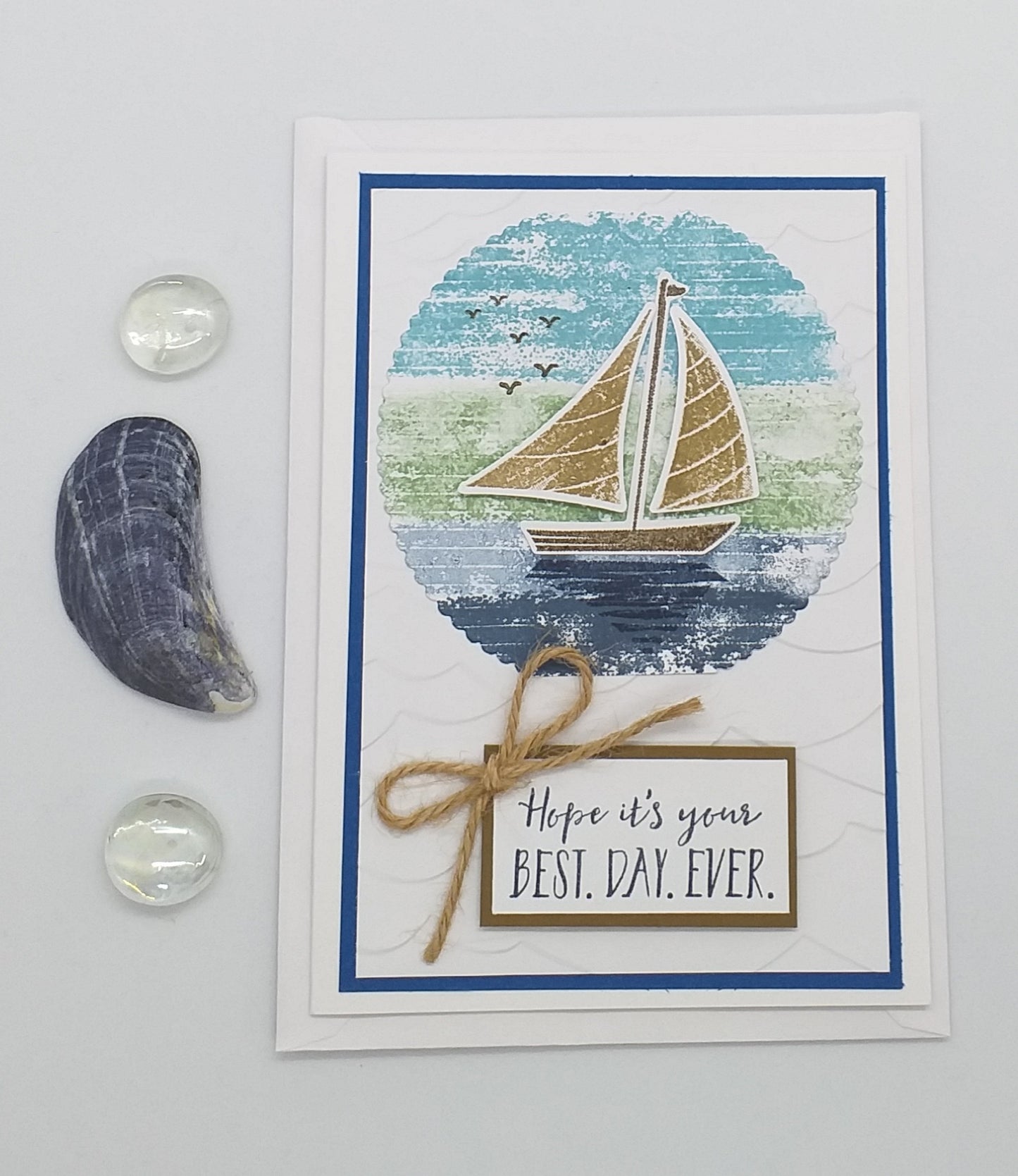 Greeting card with sailboat design and 'Hope it's your best day ever' text, placed on a beach with ocean view.