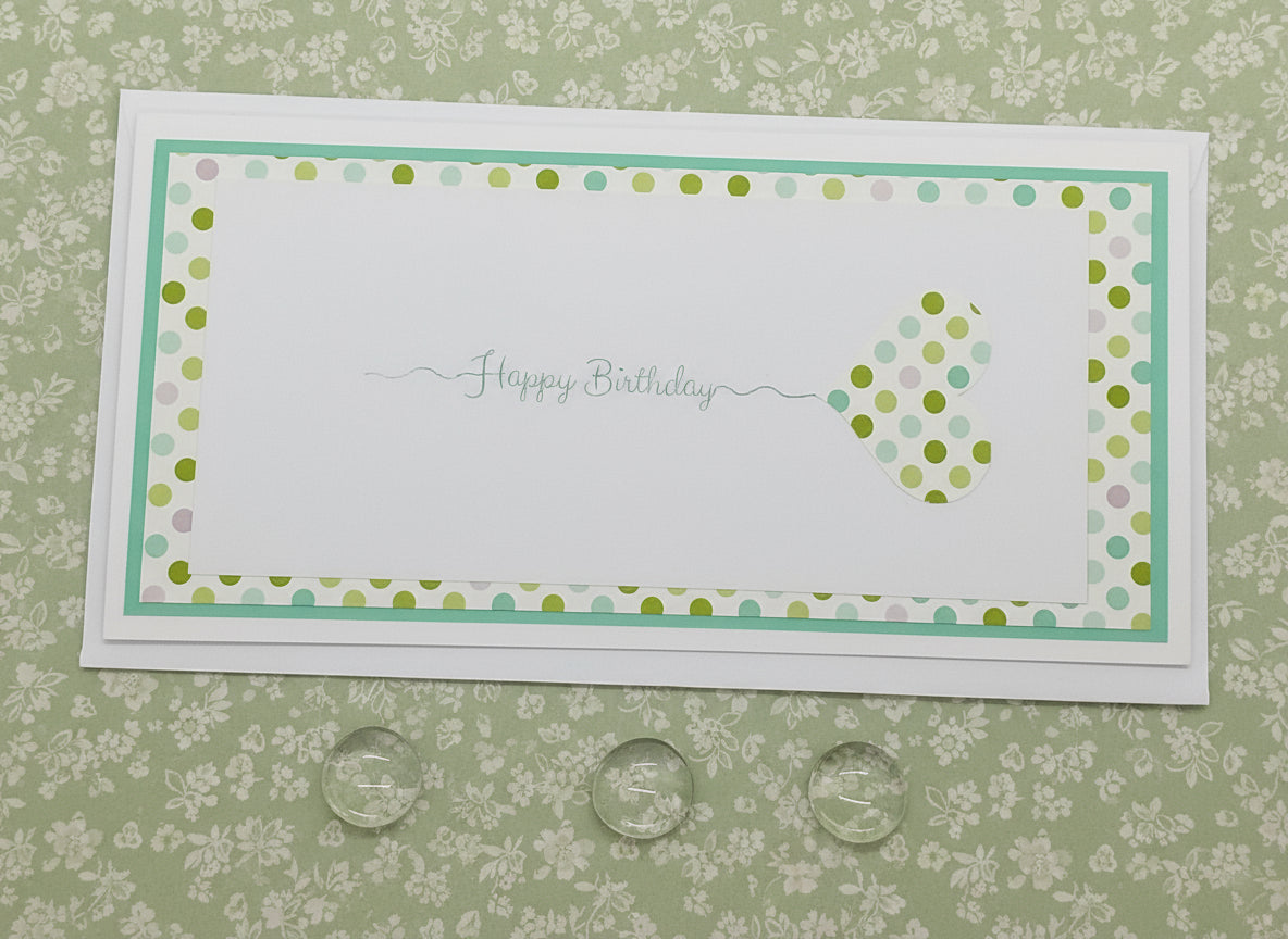 A slimline, colorful birthday card with a stamped design, featuring a white background with green and turquoise accents, and a heart-shaped graphic in the center.