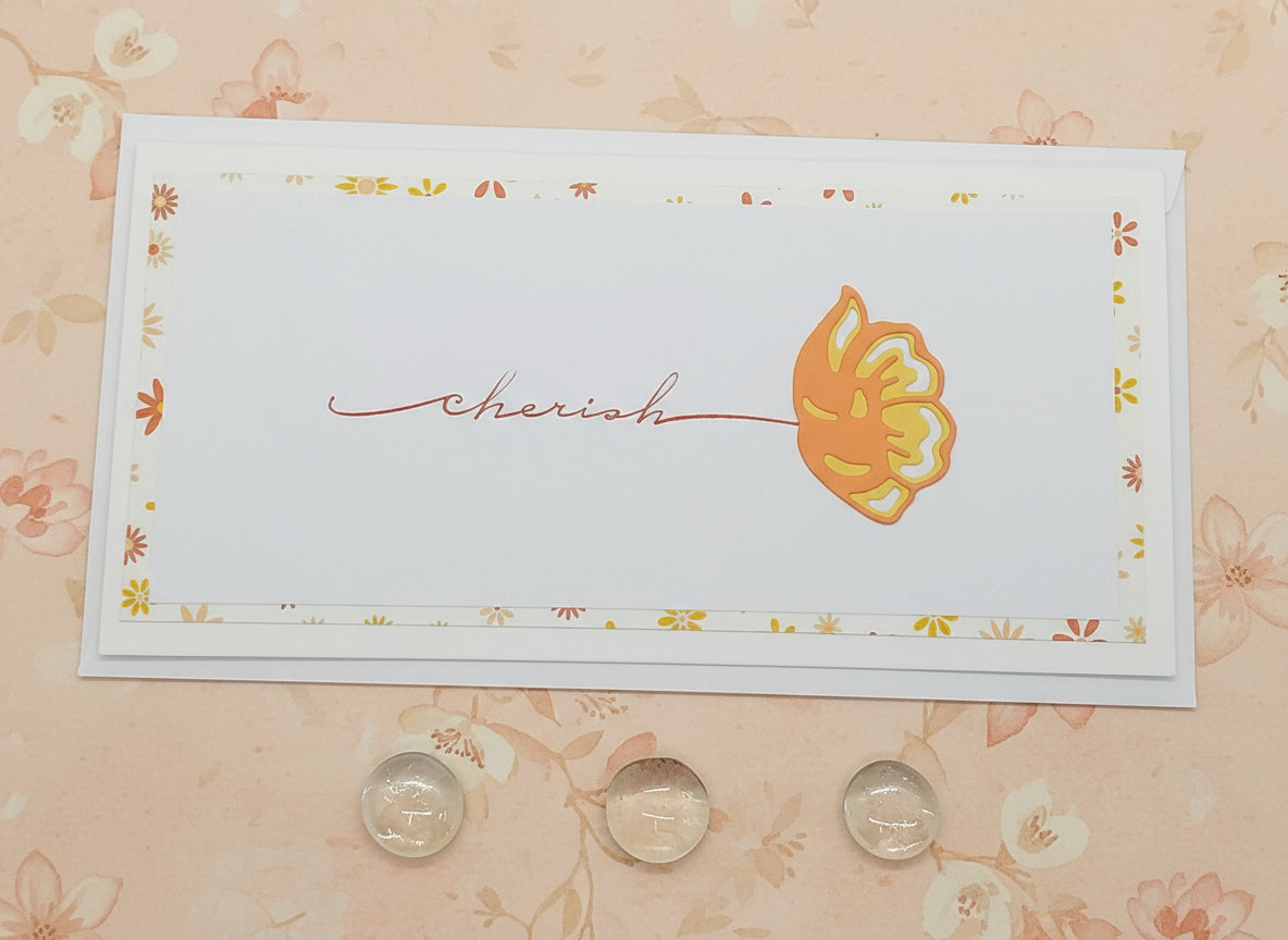 A slimline card with 'CHERISH' written in a cursive font, accompanied by a small flower cutout, on a background with a floral pattern. The card appears to be white with an orange and yellow flower design and comes with an envelope.