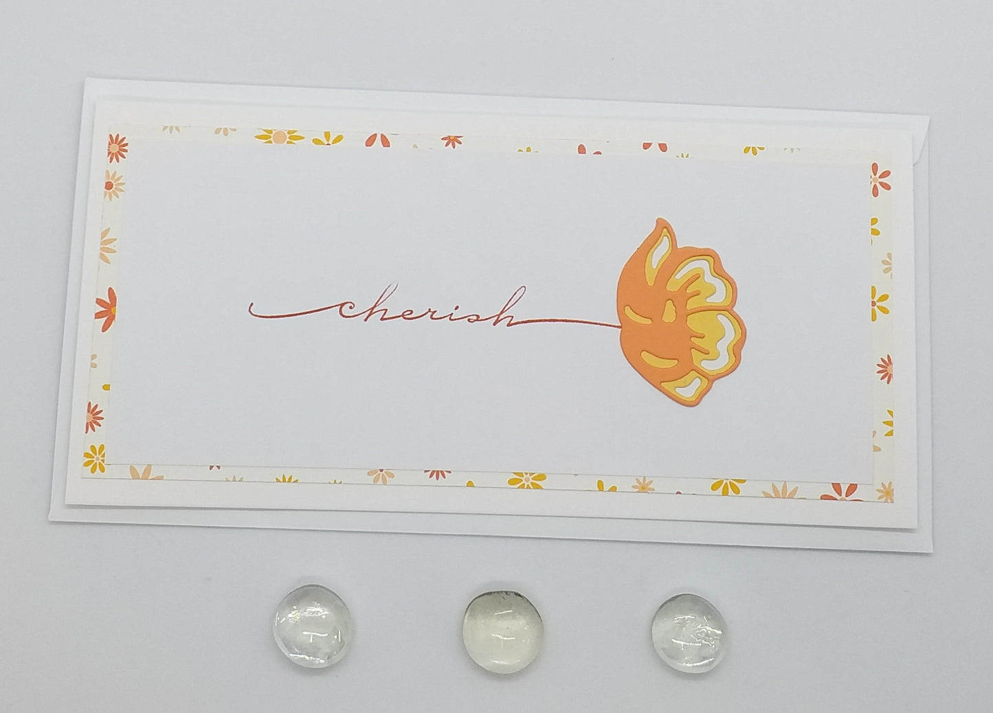 A slimline card with 'CHERISH' written in a cursive font, accompanied by a small flower cutout, on a background with a floral pattern. The card appears to be white with an orange and yellow flower design and comes with an envelope.