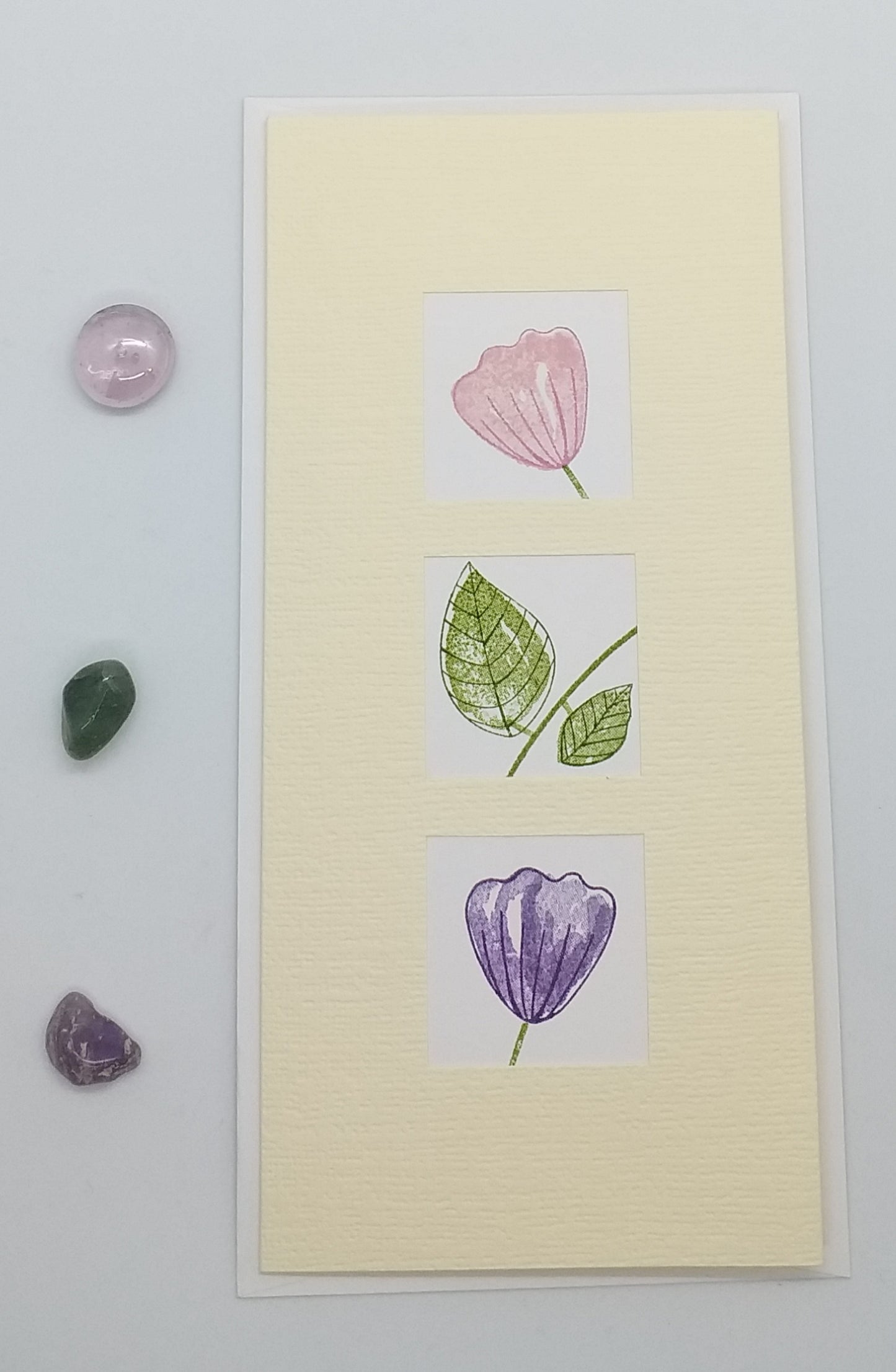 A greeting card with three windows displaying hand-stamped images of purple flowers and green leaves. The card is presented against a neutral background with small purple and green stones to the side.