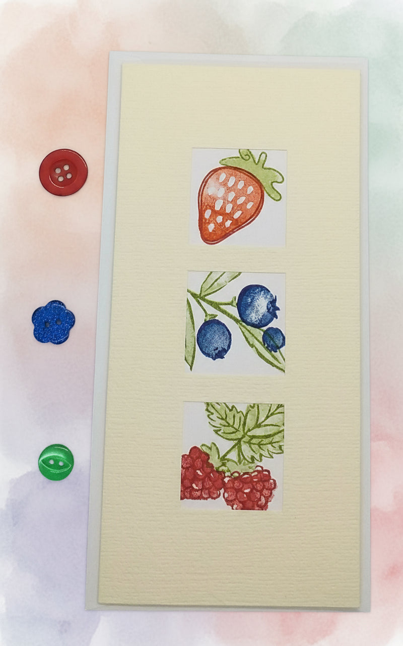 A slimline card with three windows displaying hand-stamped images of strawberries, blueberries, and raspberries, accompanied by a red and a blue button on the top edge.