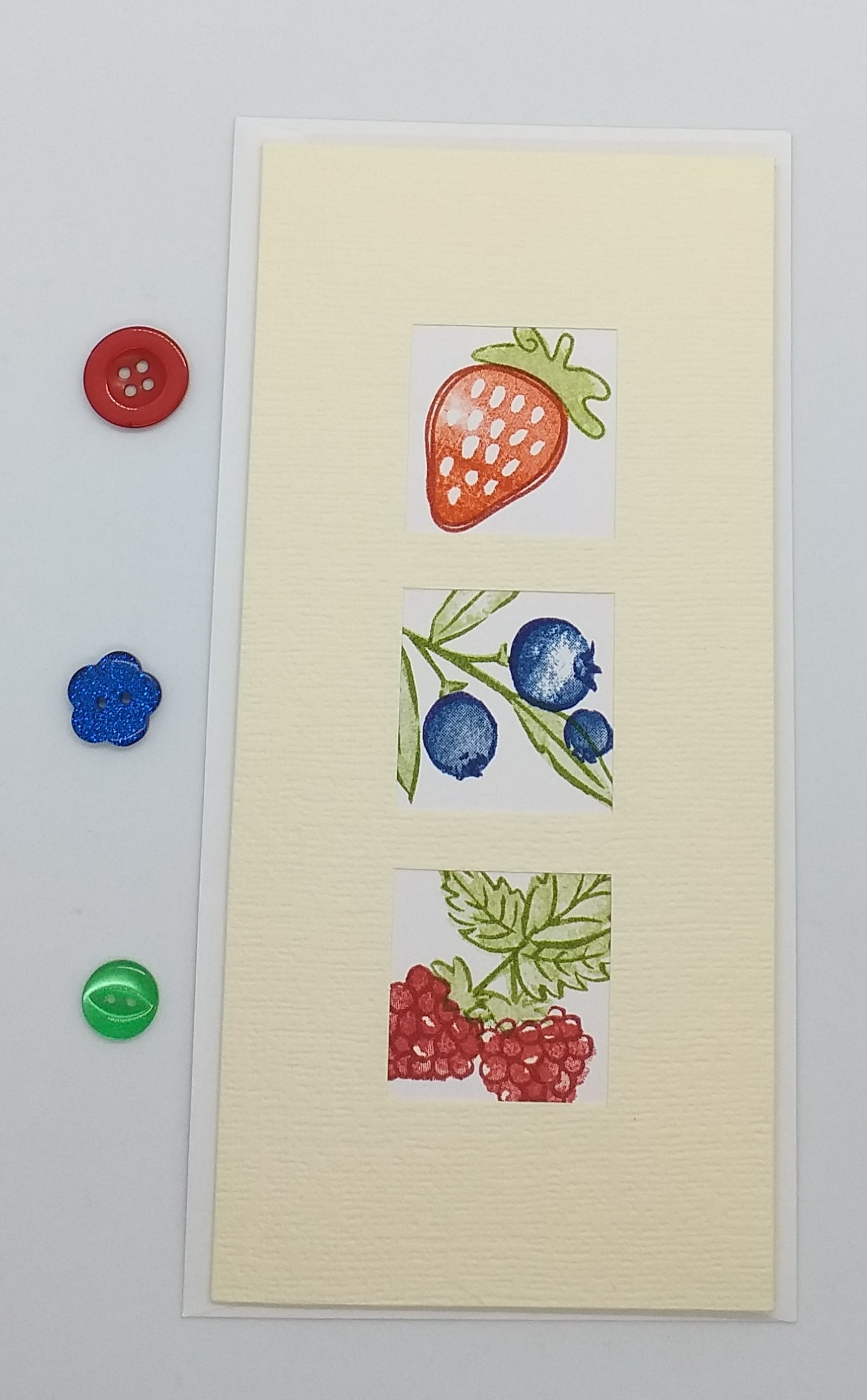 A slimline card with three windows displaying hand-stamped images of strawberries, blueberries, and raspberries, accompanied by a red and a blue button on the top edge.