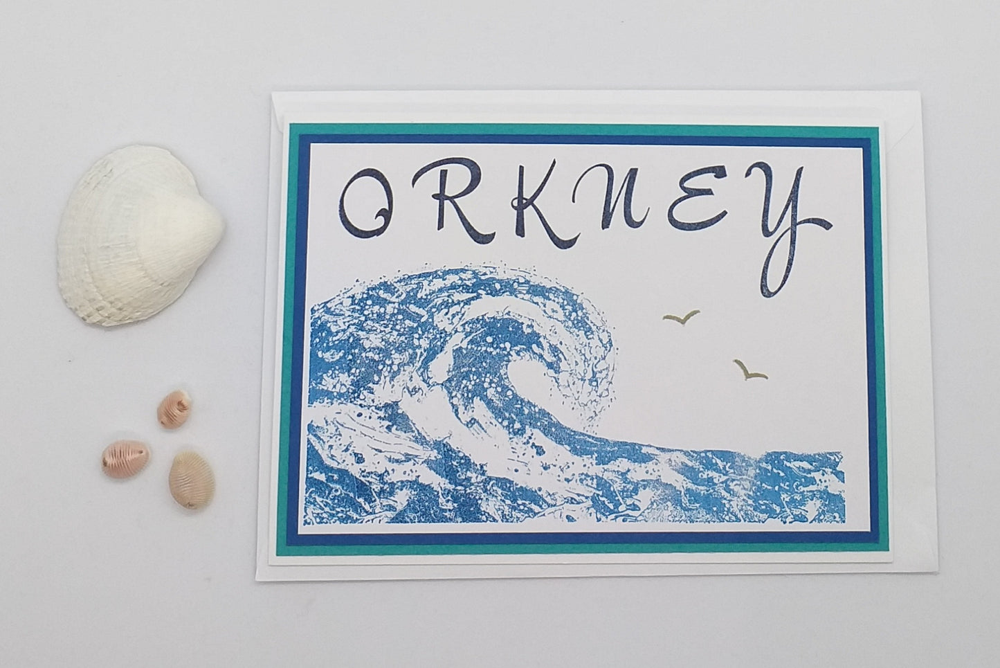 A card with a blue wave design and the word 'ORKNEY' written across the top. A small rock and a seagull figurine are also present to the left of the card.