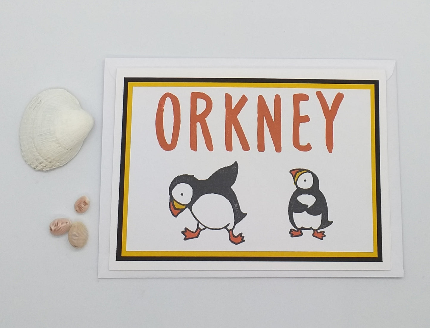 A handmade a6 greeting card with 'ORKNEY' text and illustrations of two puffins, one black and one white, on a yellow and orange background. The card is blank inside for a personal message and includes an envelope.