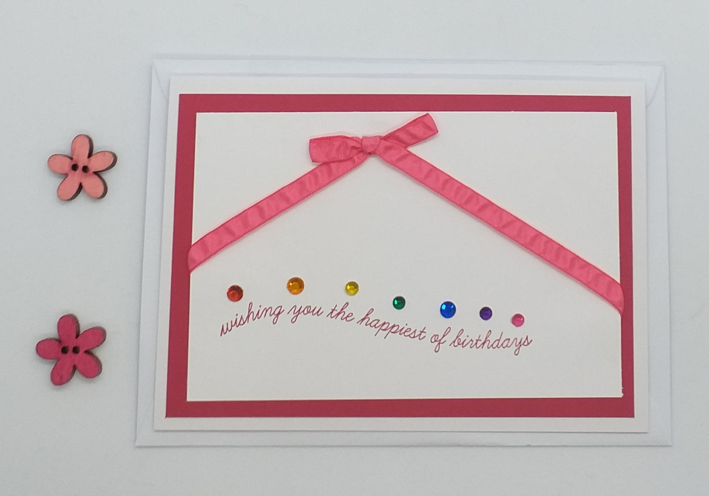 Hand-stamped birthday card with pink and pinker shades, decorated with ribbons and gems, and a written message that reads 'Wishing you the happiest of birthdays'.