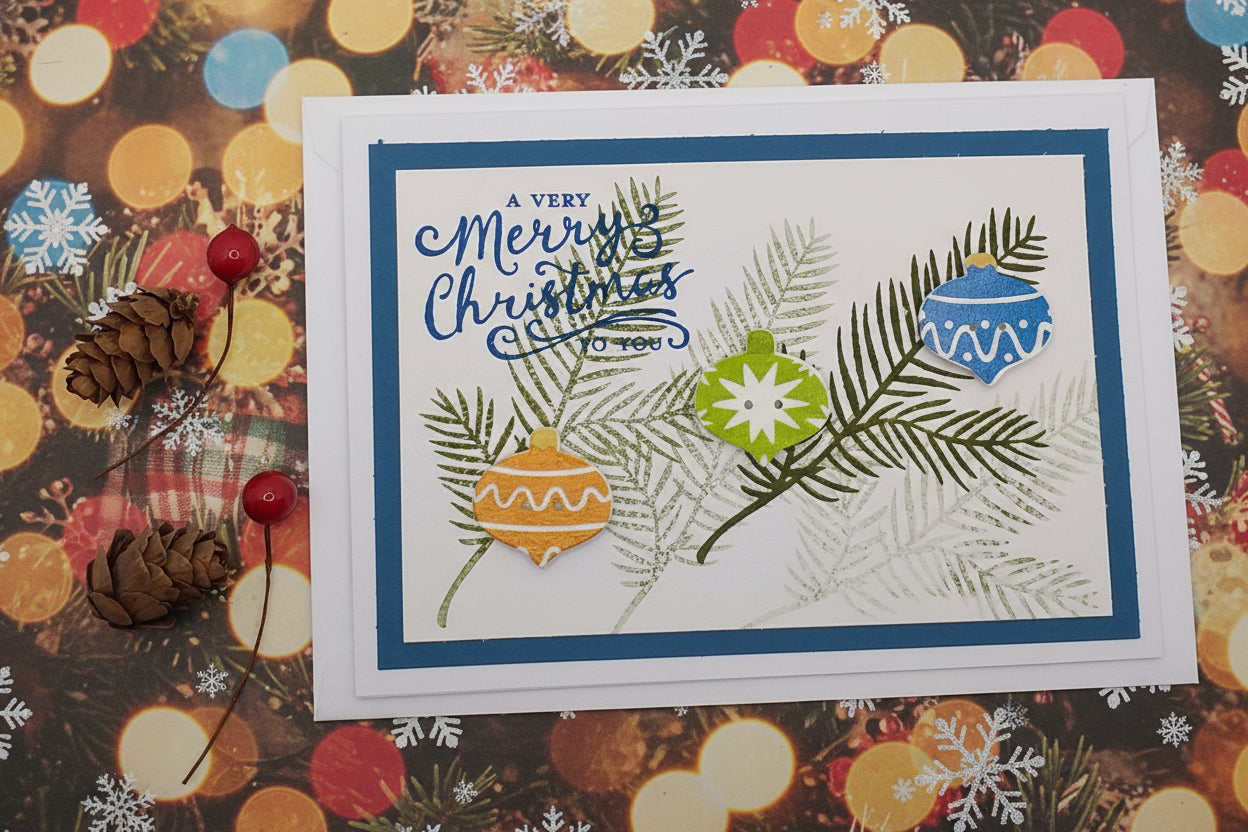 A Christmas card with 'Merry Christmas' text, decorated with three ornate bauble buttons in red, green, and yellow, and two brown leaf embellishments on a white background.