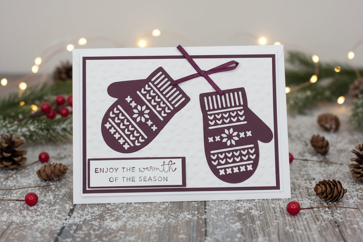 A maroon Christmas card featuring a pair of embossed mittens with design patterns, accompanied by two brown pine cones.