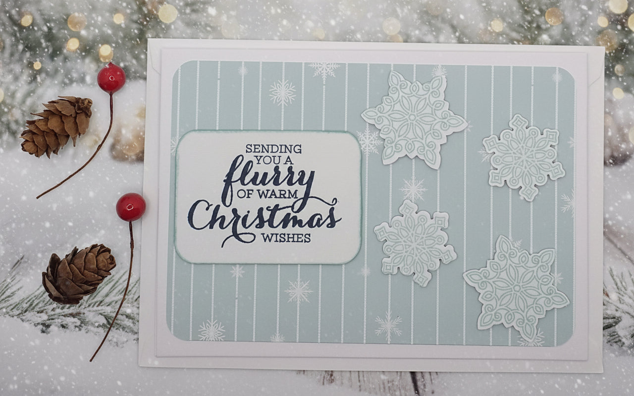 A Christmas card with 'Warming Flurries Christmas Wishes' text and decorated with snowflake shapes, accompanied by two red berries and a pine leaf embellishments.