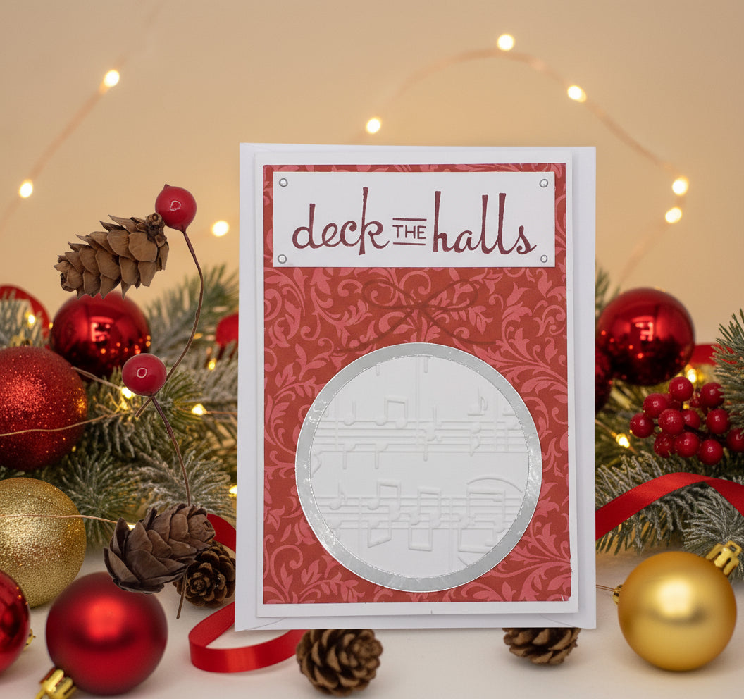A Christmas card with a red background, decorated with a musical instrument design and the text 'DECK THE HALLS'. There are two red berries and a pine cone decoration on the front.