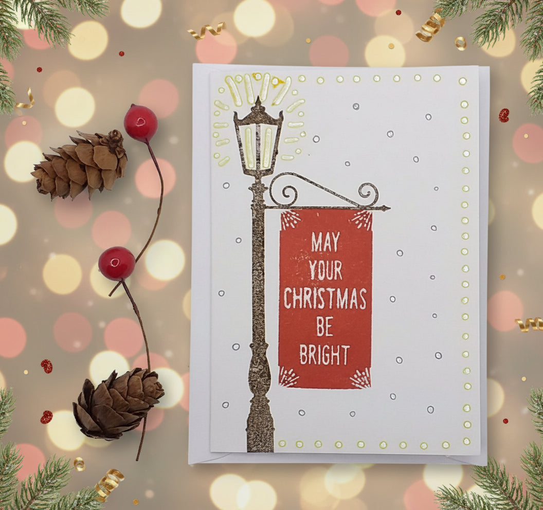 A Christmas card with a white background featuring a decorative lamp post, red banner reading 'May your Christmas be bright', and green accents, accompanied by two red berries and a pine cone decoration.