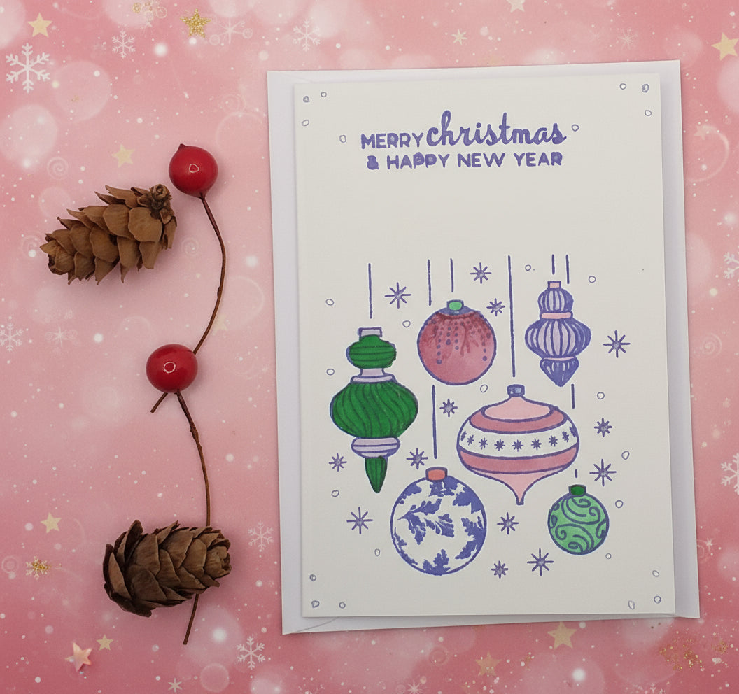 A hand-stamped and colored Christmas card depicting various Christmas ornaments, with a blank space for a personal message, accompanied by pine cones on a white background.