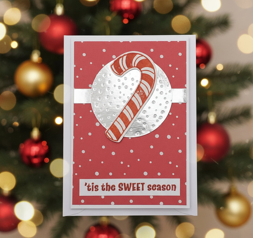 A red Christmas card with a candy cane and snowflakes design, and the text 'It's the sweet season' on the front. There are two brown pine cones to the left of the card.