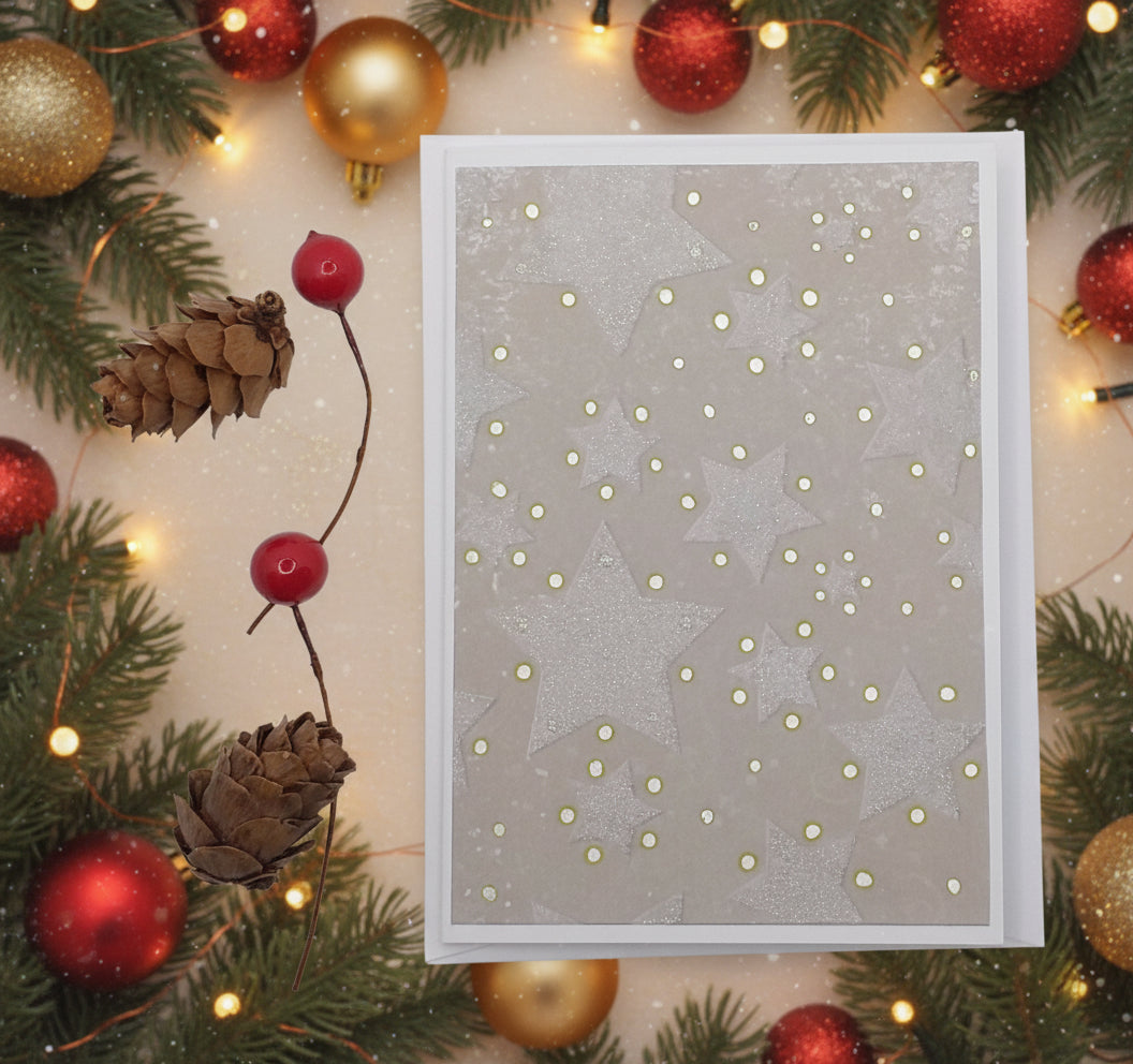 A Christmas card with stars and gold embossing, displayed alongside pine cones and a red berry, on a white background.