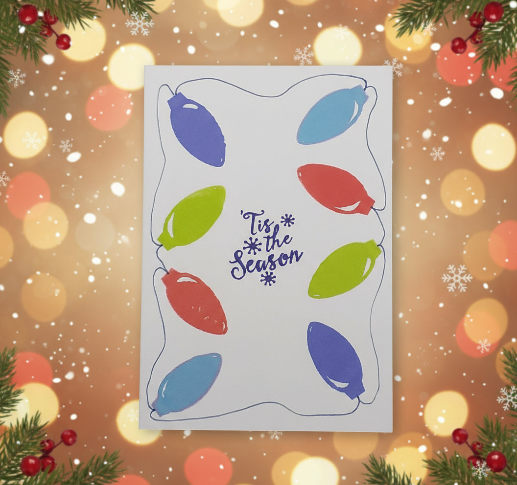 A greeting card with a blank interior, featuring a hand-stamped design of colorful Christmas lights on a white background, with the text 'tis the season' on the front. The card is displayed with a few pine cones and red berries to the side.