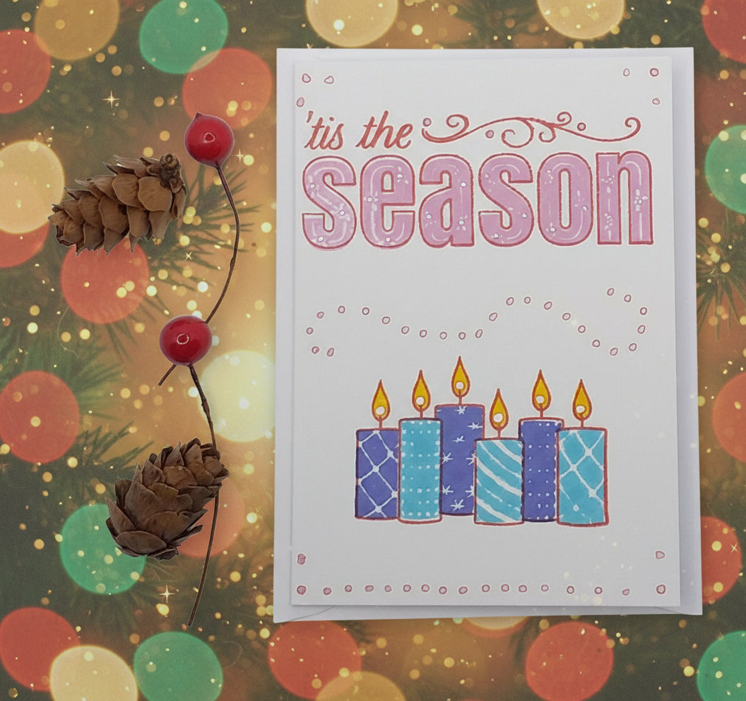 A Christmas card with 'Tis the Season' text in pink, blue candles hand-stamped in the center, accompanied by a red berry and a pine cone decoration.