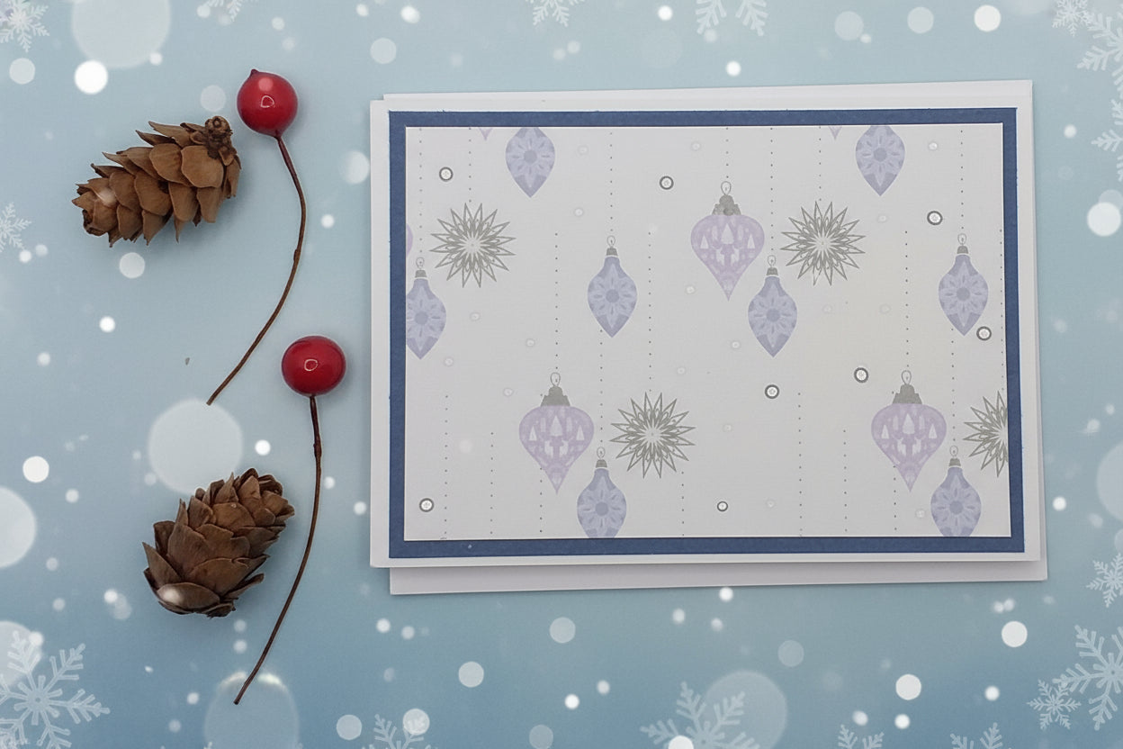 A7-sized Christmas card with a blue background featuring hanging baubles and shiny silver dots, accompanied by a pine cone and a cherry on top.