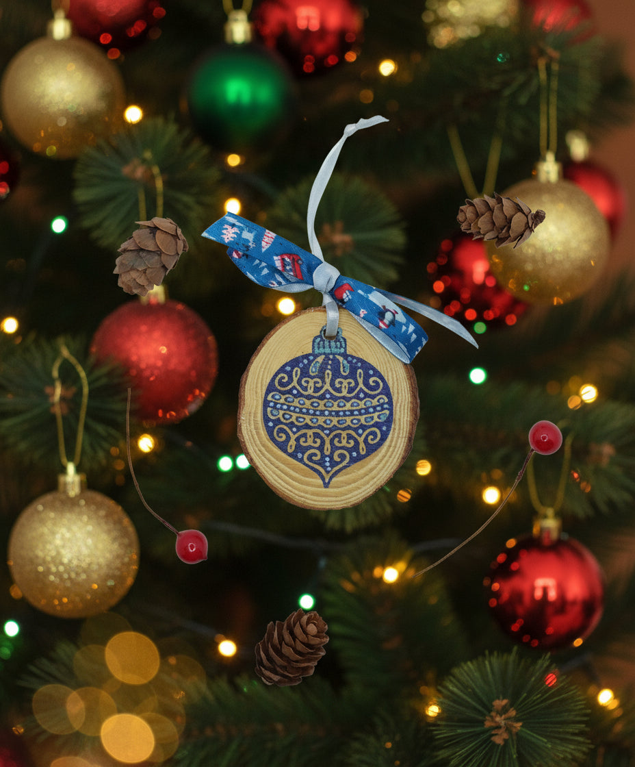 A wooden slice with a hand-stamped blue bauble design, adorned with a blue ribbon, displayed alongside pine cones and a red berry.