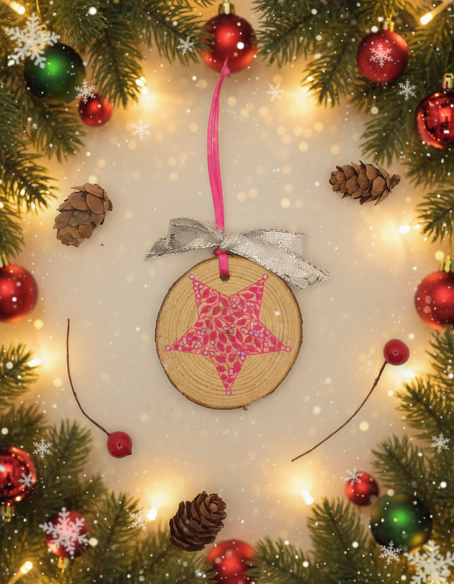 A decorative wood slice with a pink star design, adorned with a hanging ribbon and accompanied by two pine cones, against a white background.