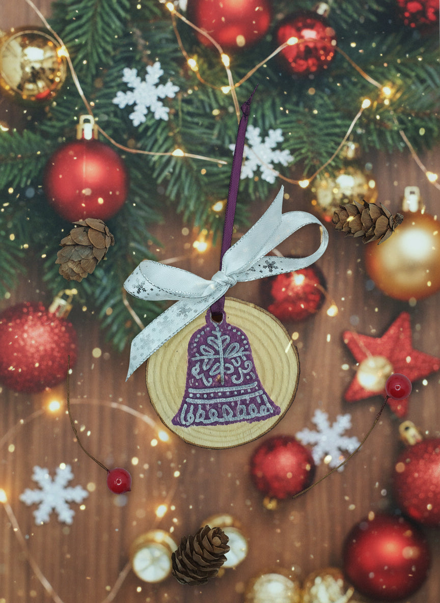 A hand-stamped, purple-colored wood slice ornament designed as a bell, with a white hanging ribbon and red berries, displayed against a white background with pine cones scattered around.