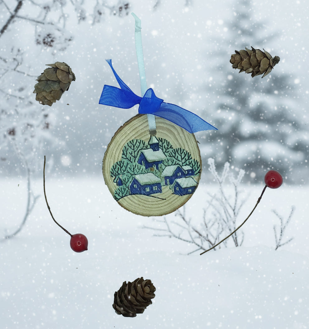 A decorative wood slice featuring a snowy village scene with a blue ribbon for hanging, accompanied by pine needles and cranberries.