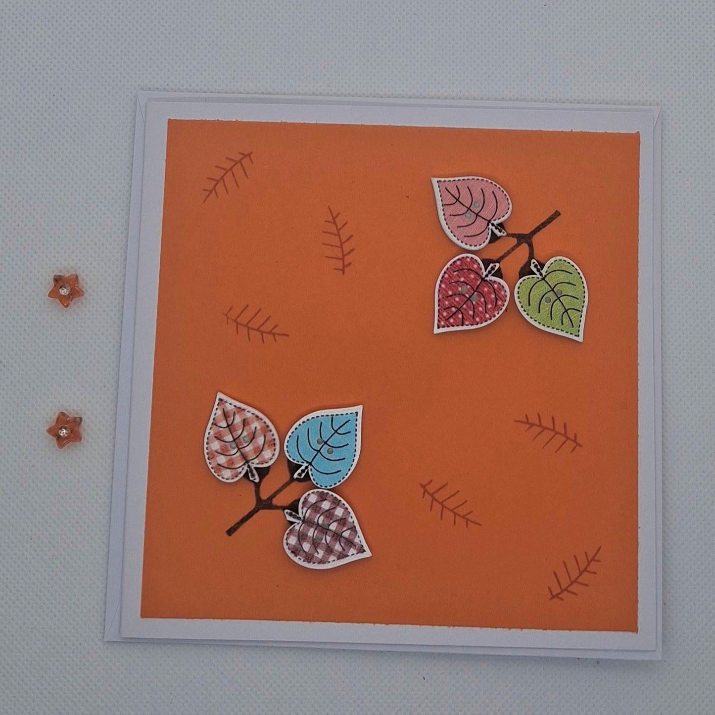 A hand-stamped button card with an orange background featuring leaf patterns and colorful buttons, displayed with a blank envelope.