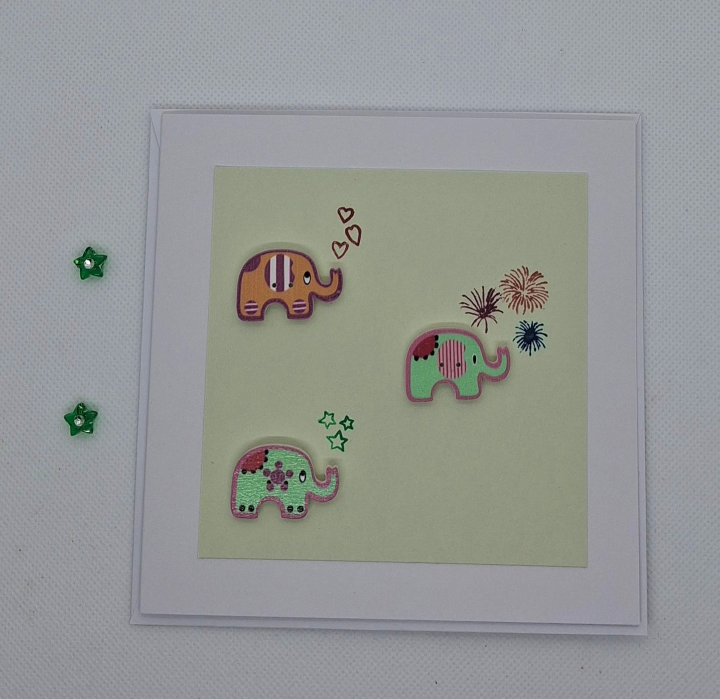 A hand-stamped and layered greeting card with three colorful elephant buttons on a green background, accompanied by small decorative elements.