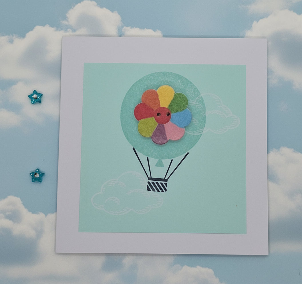 A handcrafted greeting card with a colorful balloon design at the center, featuring a button, on a blue background. The card is displayed flat with an envelope and decorative elements around it.