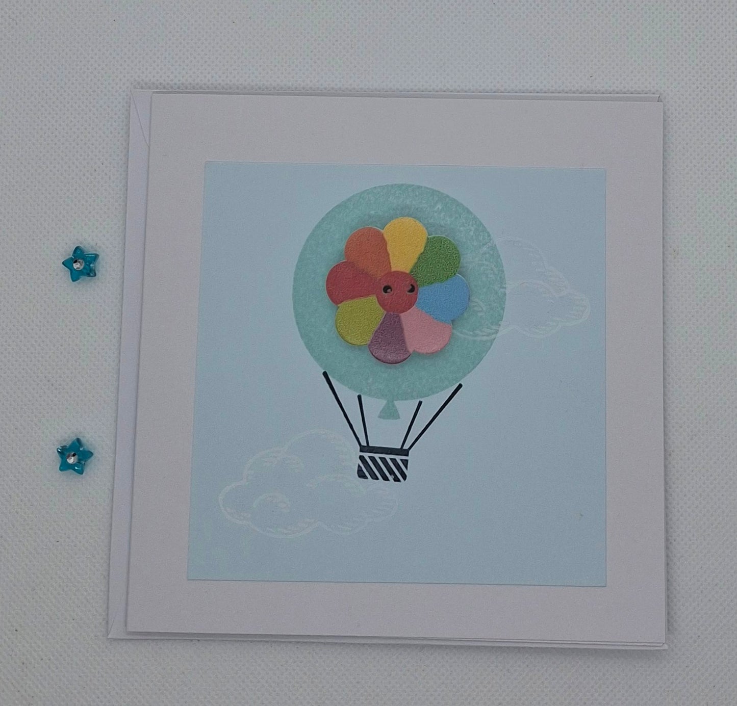 A handcrafted greeting card with a colorful balloon design at the center, featuring a button, on a blue background. The card is displayed flat with an envelope and decorative elements around it.