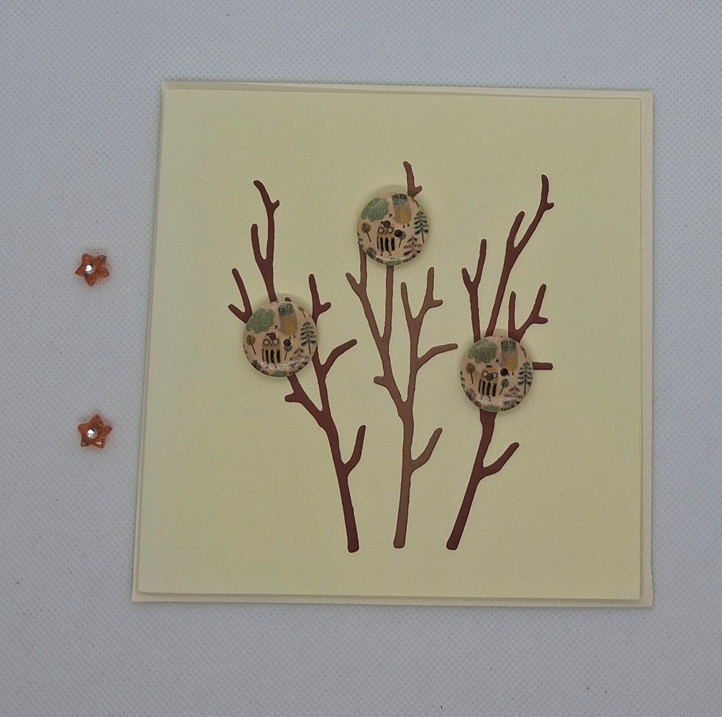 A greeting card with die-cut branches and three buttons, set against a pale background. The card is blank inside and an envelope is included.