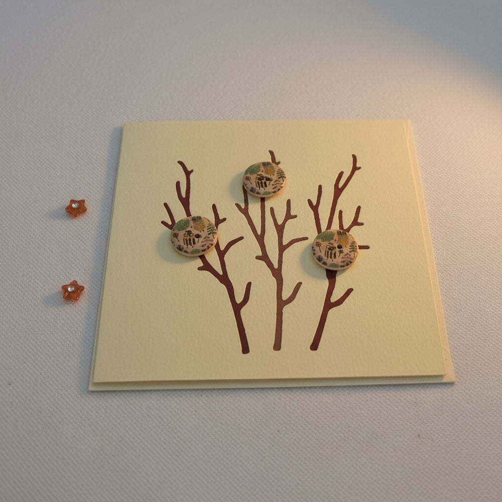 A greeting card with die-cut branches and three buttons, set against a pale background. The card is blank inside and an envelope is included.