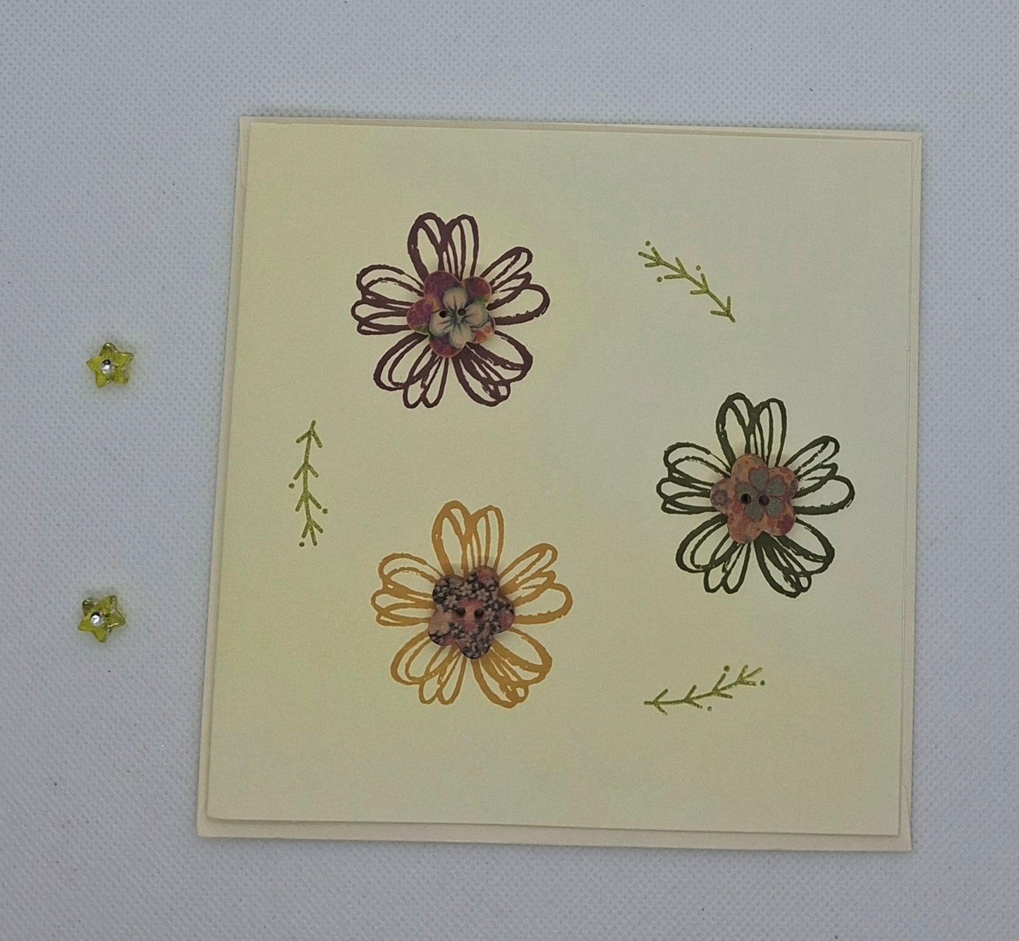 A hand stamped greeting card with three flowers and a vibrant button in the center of each flower. The card is 13.