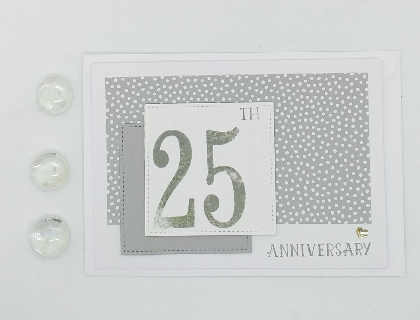 A hand-stamped 25th anniversary card with multiple layers, featuring a '25' numeral, dots and a heart embellishment, accompanied by small beads.