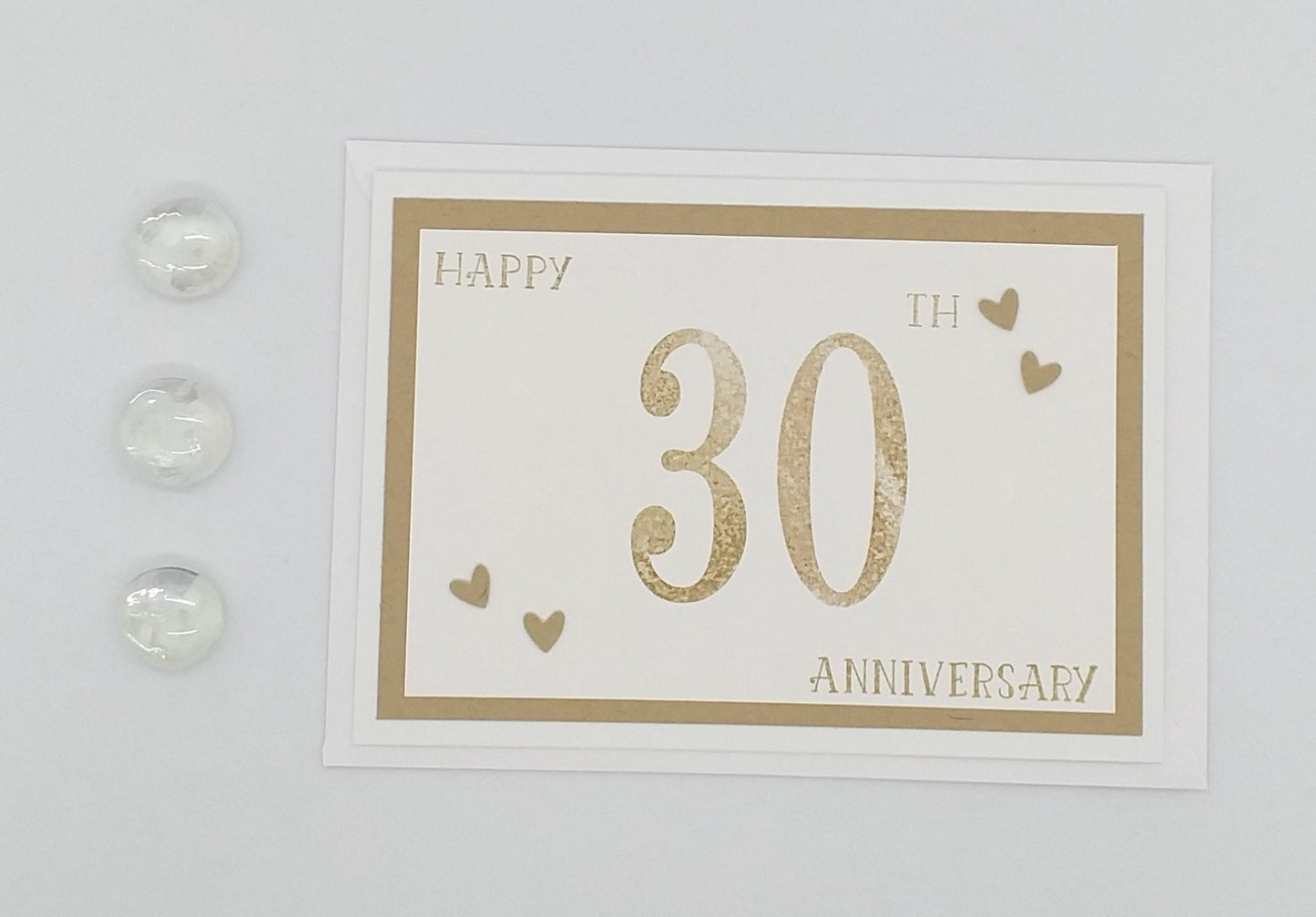 A greeting card with 'Happy 30th Anniversary' text in gold, surrounded by small hearts and a stamped gold number '30' on a white background. Two small clear beads are also visible on the card.