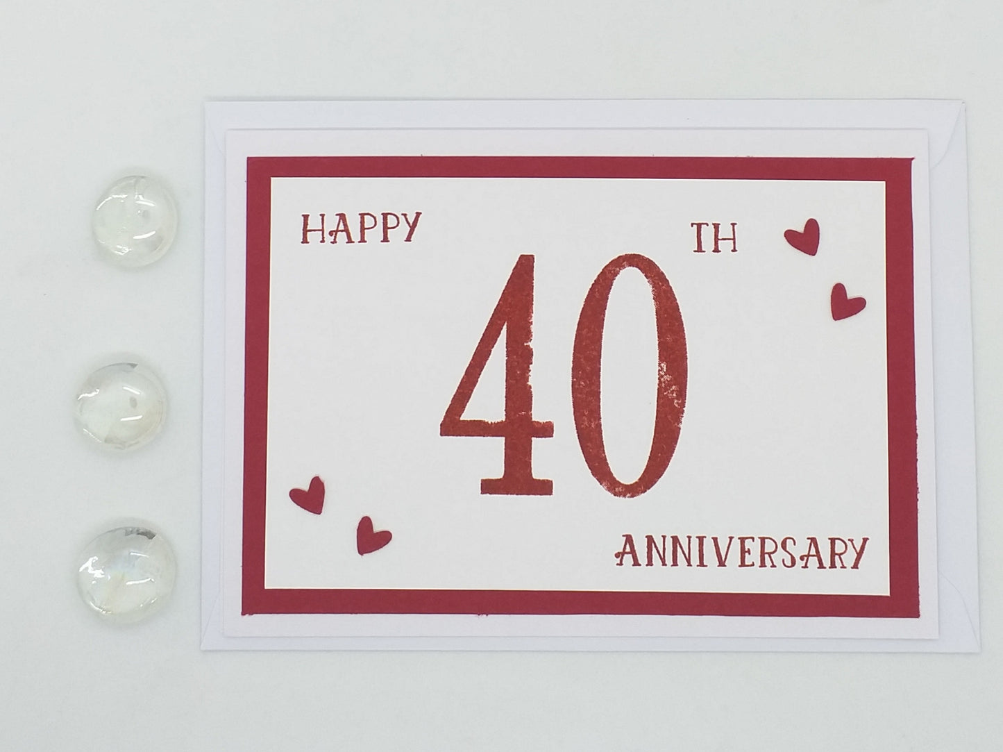 A hand stamped anniversary card with 'HAPPY 40TH ANNIVERSARY' written in bold red letters, surrounded by small red hearts on the corners. The card is placed on a white surface with three clear gemstones to the side.