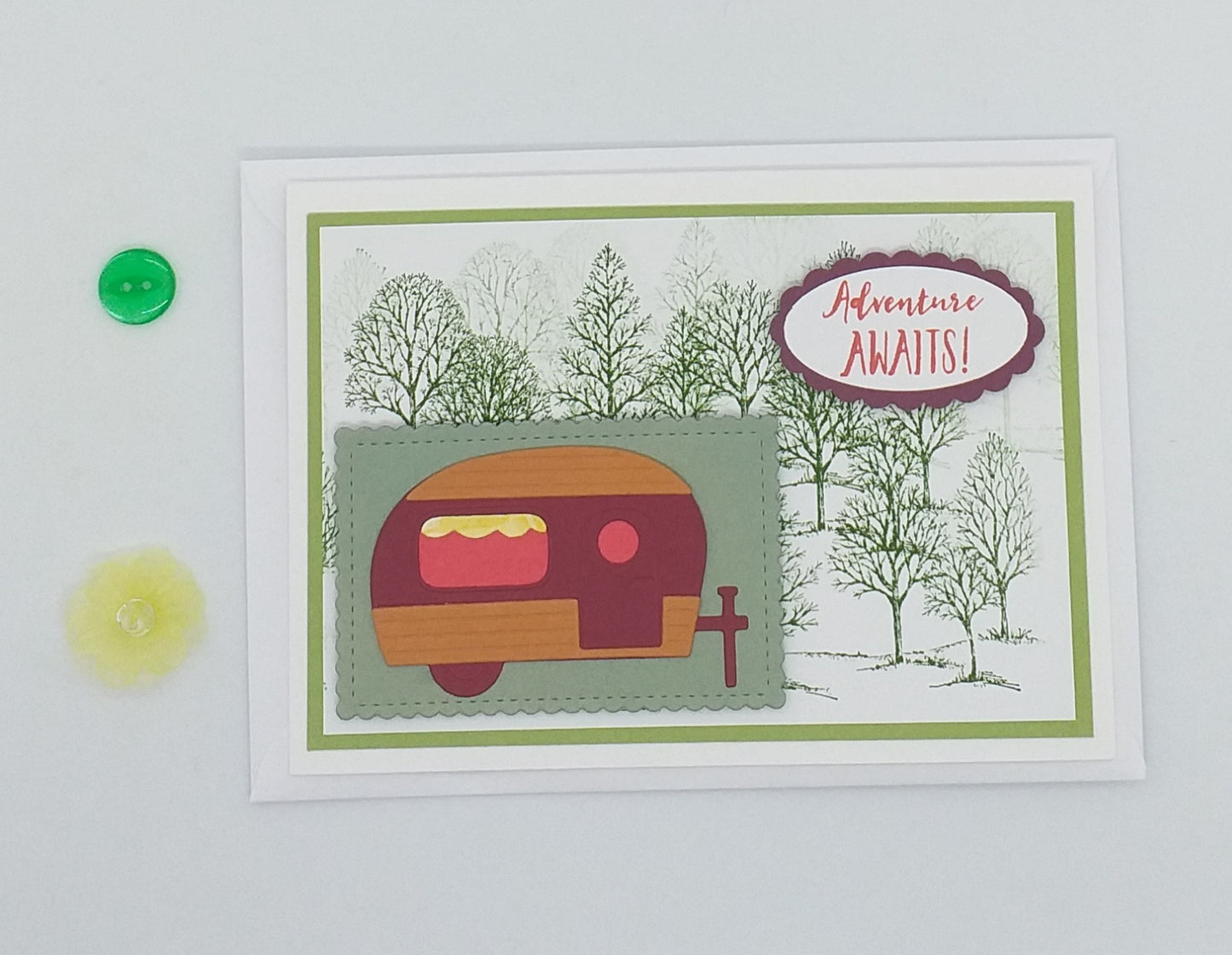 A greeting card with a colorful design featuring a trailer and trees, titled 'Adventure Awaits!' and a small green object to the side.
