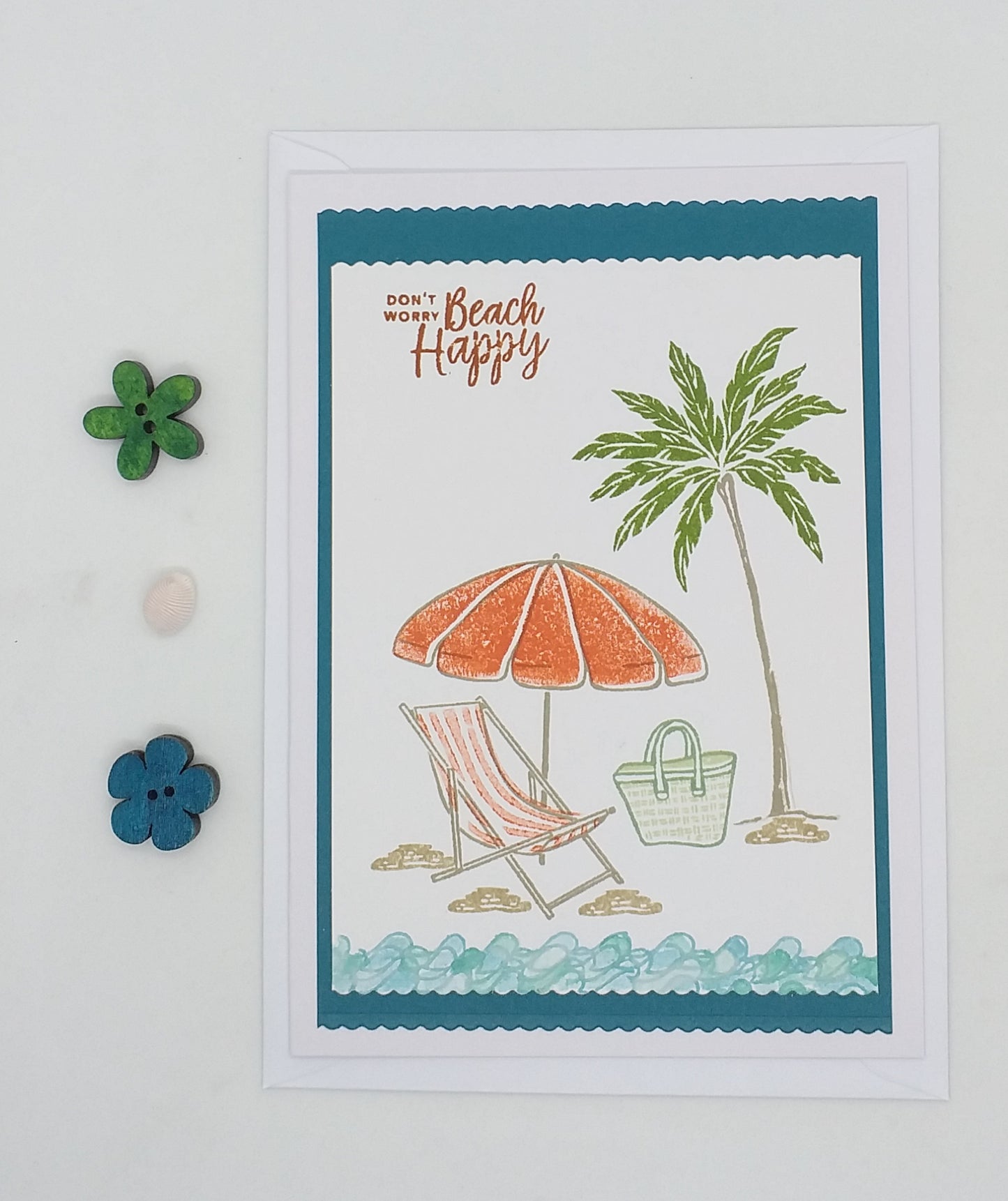 A greeting card with a beach scene illustration, including a chair, umbrella, and palm tree. The text 'Beach Happy' is featured on the card, which is placed on a white surface with decorative buttons around it.