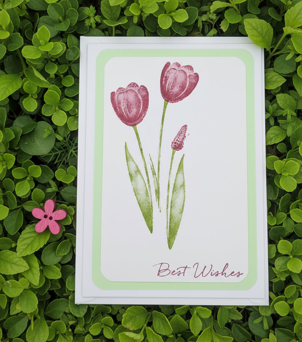 A greeting card with an envelope, featuring a hand-stamped design of tulips in shades of pink and green on a white background, with the words 'Best Wishes' written at the bottom.